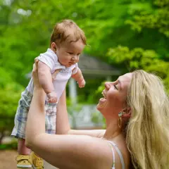 Szczęśliwa mama podrzuca niemowlaka na tle zieleni. Nawet jeśli maluch ma katar, radość z zabawy jest bezcenna.