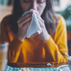 Kobieta w żółtym swetrze dmucha nos w chusteczkę, obok kubek z herbatą i tabletki.