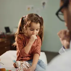 Mała dziewczynka z długimi włosami, siedząca na kozetce lekarskiej, kaszle, trzymając dłoń przy ustach. Długotrwały kaszel u dziecka bez zmian osłuchowych to problem, który martwi rodziców.