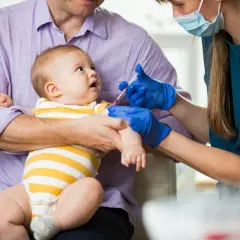 Ojciec trzyma dziecko, które otrzymuje szczepienie przeciwko pneumokokom. Lekarka w rękawiczkach podaje zastrzyk.
