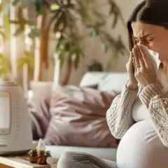 Ciężarna kobieta z chusteczką w dłoniach, siedząca na kanapie obok nawilżacza powietrza.