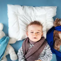Mały chłopiec z suchym kaszlem u niemowlaka, otulony szalikiem, leży obok pluszowych zabawek.