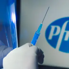 Ręka w rękawiczce trzyma strzykawkę z niebieskim płynem na tle logo Pfizer. Dyskusja o skutkach ubocznych szczepionki Pfizer trwa.