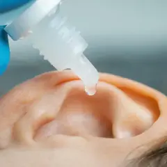 Kropla leku spada do ucha, które trzyma ręka w niebieskiej rękawiczce.