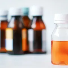 Pomarańczowy syrop na rozrzedzenie wydzieliny dla dzieci w buteleczce. W tle inne leki.