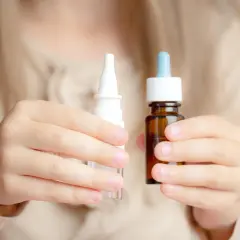 Dziecko trzyma w dłoniach dwa opakowania z kroplami do nosa: biały spray i brązową buteleczkę z pipetą.