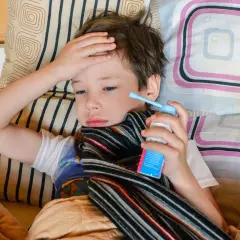 Chory chłopiec leży w łóżku, trzymając w dłoni inhalator. Jego ręka spoczywa na czole, co może sugerować gorączkę lub ból głowy, jedne z objawów astmy u dzieci.