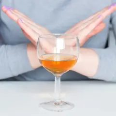 Dłonie skrzyżowane na znak stopu nad kieliszkiem z bursztynowym płynem. To symbol uczulenia na alkohol.