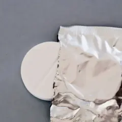 Biała tabletka z nacięciem obok srebrnego opakowania na szarym tle.