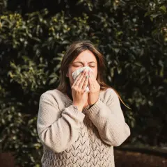 Kobieta w beżowym swetrze chusteczką wyciera nos na tle zielonych liści. Wiosna, na co teraz alergia?