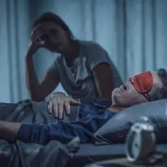 Zestresowana kobieta szuka sposobów na chrapanie swojego partnera, który głośno śpi w czerwonej opasce na oczy.