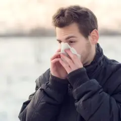 Młody mężczyzna w zimowej kurtce chowa twarz w chusteczkę, cierpiąc na przewlekły mokry kaszel.