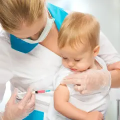 Pielęgniarka podaje niemowlęciu szczepionkę przeciwko meningokokom. Dziecko spokojnie siedzi na rękach.