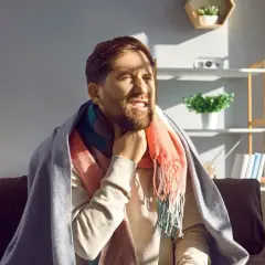 Mężczyzna z bólem gardła, otulony kocem i szalikiem, siedzi na kanapie.