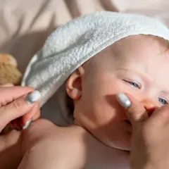 Niebieskooki noworodek w białej sapce, delikatnie dotykany przez dłoń z błyszczącymi paznokciami. Obok leży drewniana szczoteczka.