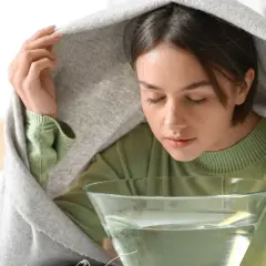 Kobieta pochyla się nad miską z wodą, wdychając parę pod kocem.