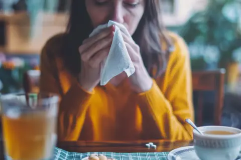 Kobieta w żółtym swetrze dmucha nos w chusteczkę, obok kubek z herbatą i tabletki.