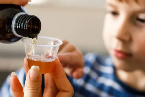 Dziecko patrzy, jak dorosły nalewa syrop z butelki do miarki.