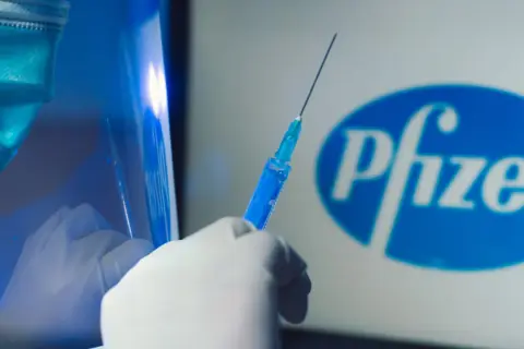 Ręka w rękawiczce trzyma strzykawkę z niebieskim płynem na tle logo Pfizer. Dyskusja o skutkach ubocznych szczepionki Pfizer trwa.