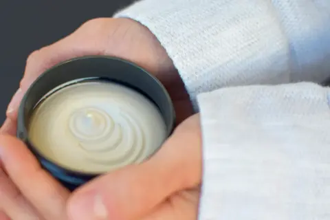 Dłonie trzymające słoiczek z białą, kremową maścią o spiralnym wzorze.