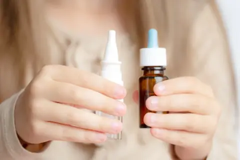 Dziecko trzyma w dłoniach dwa opakowania z kroplami do nosa: biały spray i brązową buteleczkę z pipetą.