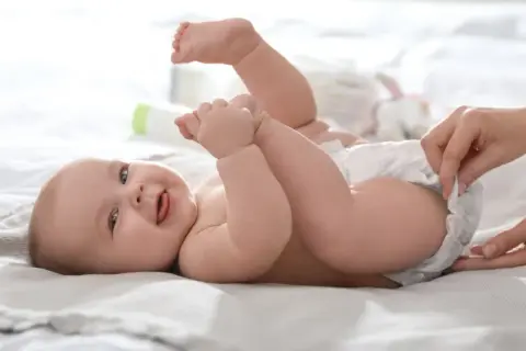 Uśmiechnięte dziecko leży na plecach, a ręka mamy zmienia mu pieluszkę, zapobiegając pieluszkowemu zapaleniu skóry.