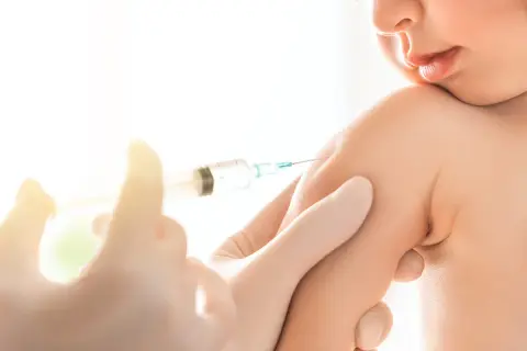 Małe dziecko otrzymuje szczepionkę 6w1. Ręka w rękawiczce trzyma strzykawkę, podając zastrzyk w ramię malucha.