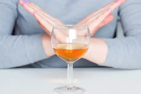 Dłonie skrzyżowane na znak stopu nad kieliszkiem z bursztynowym płynem. To symbol uczulenia na alkohol.