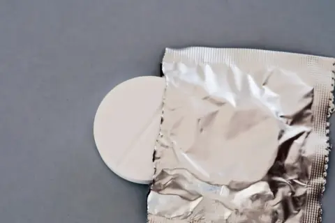 Biała tabletka z nacięciem obok srebrnego opakowania na szarym tle.