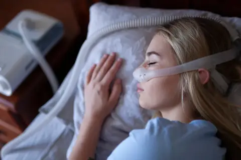 Kobieta śpi z maską CPAP na twarzy, obok stoi aparat do terapii bezdechu sennego.