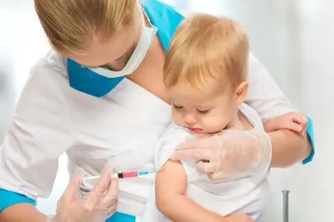 Pielęgniarka podaje niemowlęciu szczepionkę przeciwko meningokokom. Dziecko spokojnie siedzi na rękach.