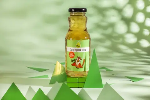 Butelka syropu z pędów sosny na zielonym tle, otoczona papierowymi choinkami.