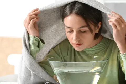 Kobieta pochyla się nad miską z wodą, wdychając parę pod kocem.