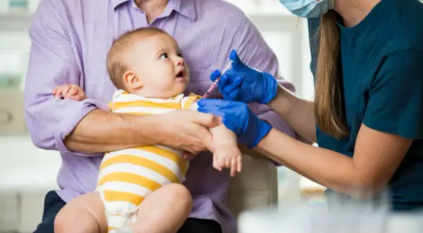 Ojciec trzyma dziecko, które otrzymuje szczepienie przeciwko pneumokokom. Lekarka w rękawiczkach podaje zastrzyk.