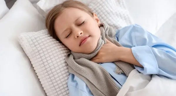 Dziewczynka w niebieskiej piżamie i szaliku trzyma się za bolące gardło, leżąc w łóżku.