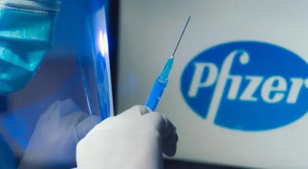 Ręka w rękawiczce trzyma strzykawkę z niebieskim płynem na tle logo Pfizer. Dyskusja o skutkach ubocznych szczepionki Pfizer trwa.