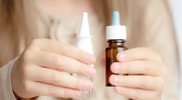 Dziecko trzyma w dłoniach dwa opakowania z kroplami do nosa: biały spray i brązową buteleczkę z pipetą.