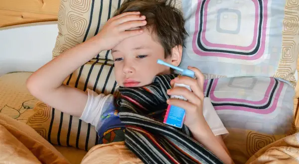Chory chłopiec leży w łóżku, trzymając w dłoni inhalator. Jego ręka spoczywa na czole, co może sugerować gorączkę lub ból głowy, jedne z objawów astmy u dzieci.