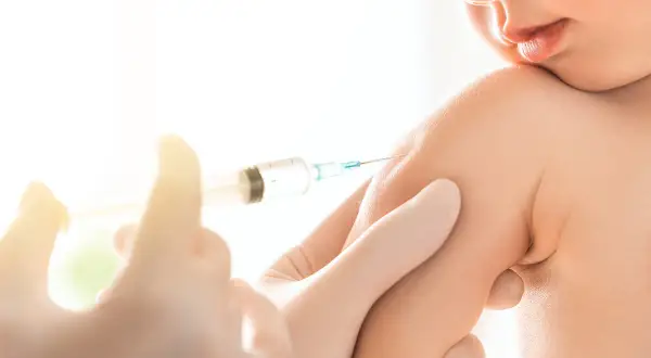 Małe dziecko otrzymuje szczepionkę 6w1. Ręka w rękawiczce trzyma strzykawkę, podając zastrzyk w ramię malucha.