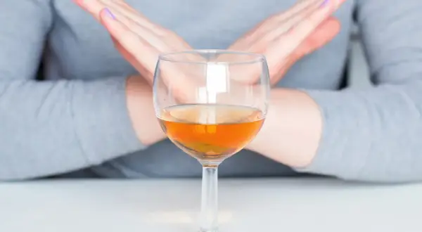 Dłonie skrzyżowane na znak stopu nad kieliszkiem z bursztynowym płynem. To symbol uczulenia na alkohol.
