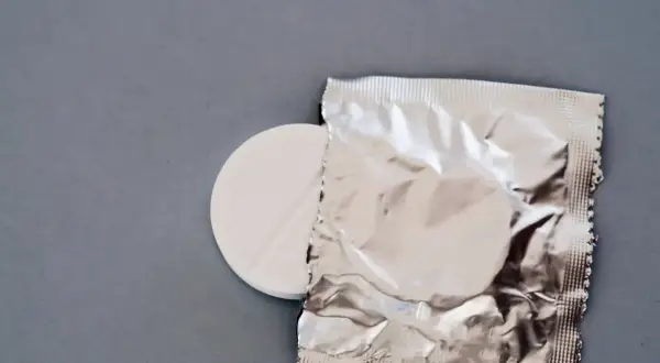 Biała tabletka z nacięciem obok srebrnego opakowania na szarym tle.