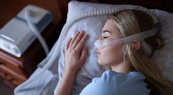 Kobieta śpi z maską CPAP na twarzy, obok stoi aparat do terapii bezdechu sennego.