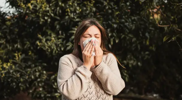 Kobieta w beżowym swetrze chusteczką wyciera nos na tle zielonych liści. Wiosna, na co teraz alergia?