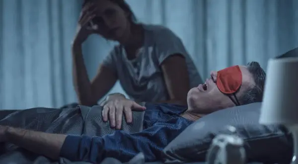 Zestresowana kobieta szuka sposobów na chrapanie swojego partnera, który głośno śpi w czerwonej opasce na oczy.