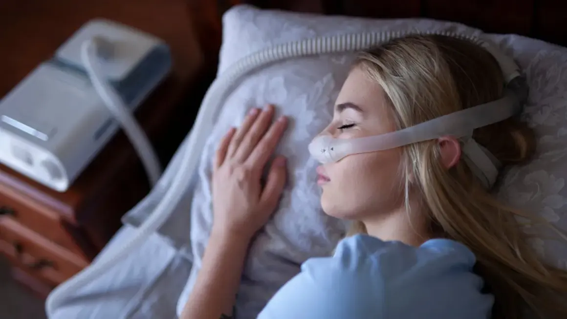 Kobieta śpi z maską CPAP na twarzy, obok stoi aparat do terapii bezdechu sennego.