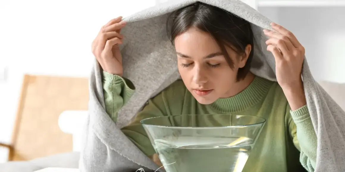 Kobieta pochyla się nad miską z wodą, wdychając parę pod kocem.