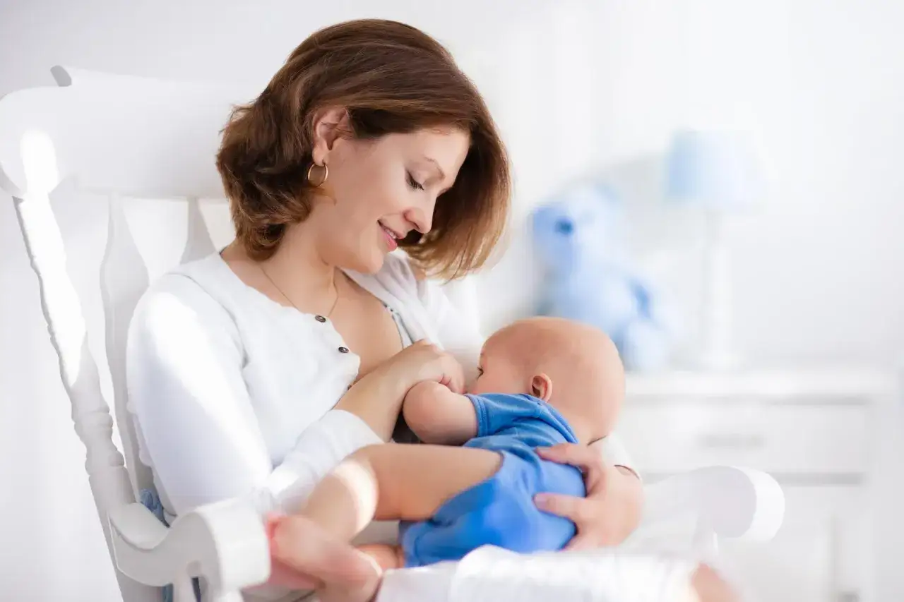 Uśmiechnięta mama karmi piersią dziecko w bujanym fotelu. Nawet jeśli maluch ma refluks, chwile bliskości są bezcenne.