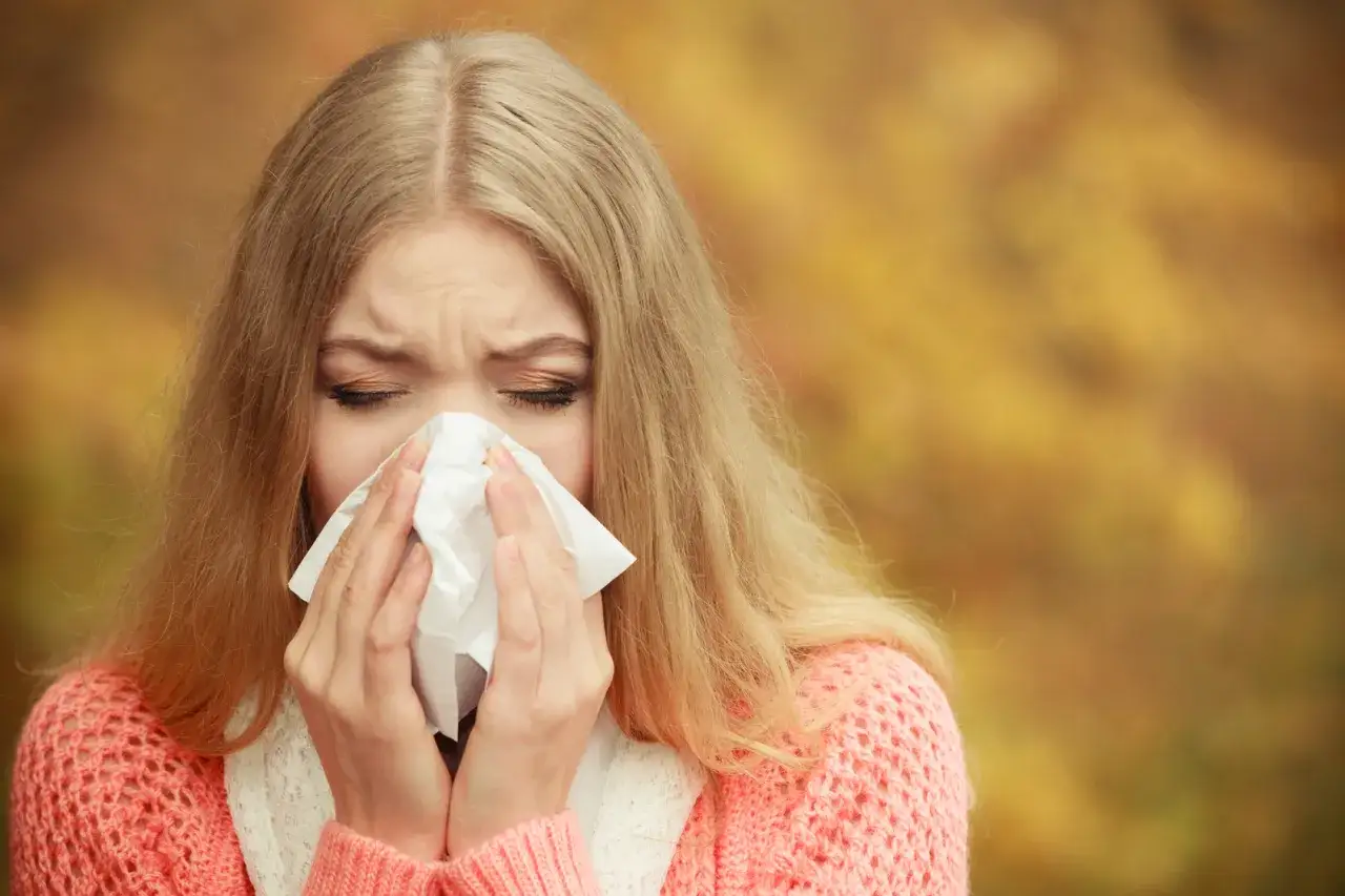 Blondynka z chusteczką w dłoniach, zamyślona, z nosem czerwonym od kataru. Jesień, liście na drzewach. Czy to na co teraz alergia?