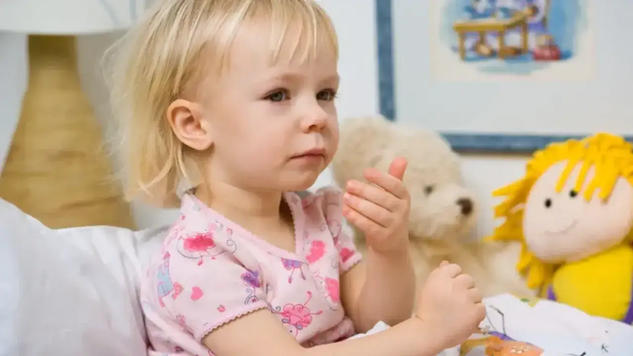 Mała dziewczynka z blond włosami, w różowej piżamce, siedzi w łóżku z pluszakami. Martwi się długotrwałym kaszlem u dziecka bez zmian osłuchowych.