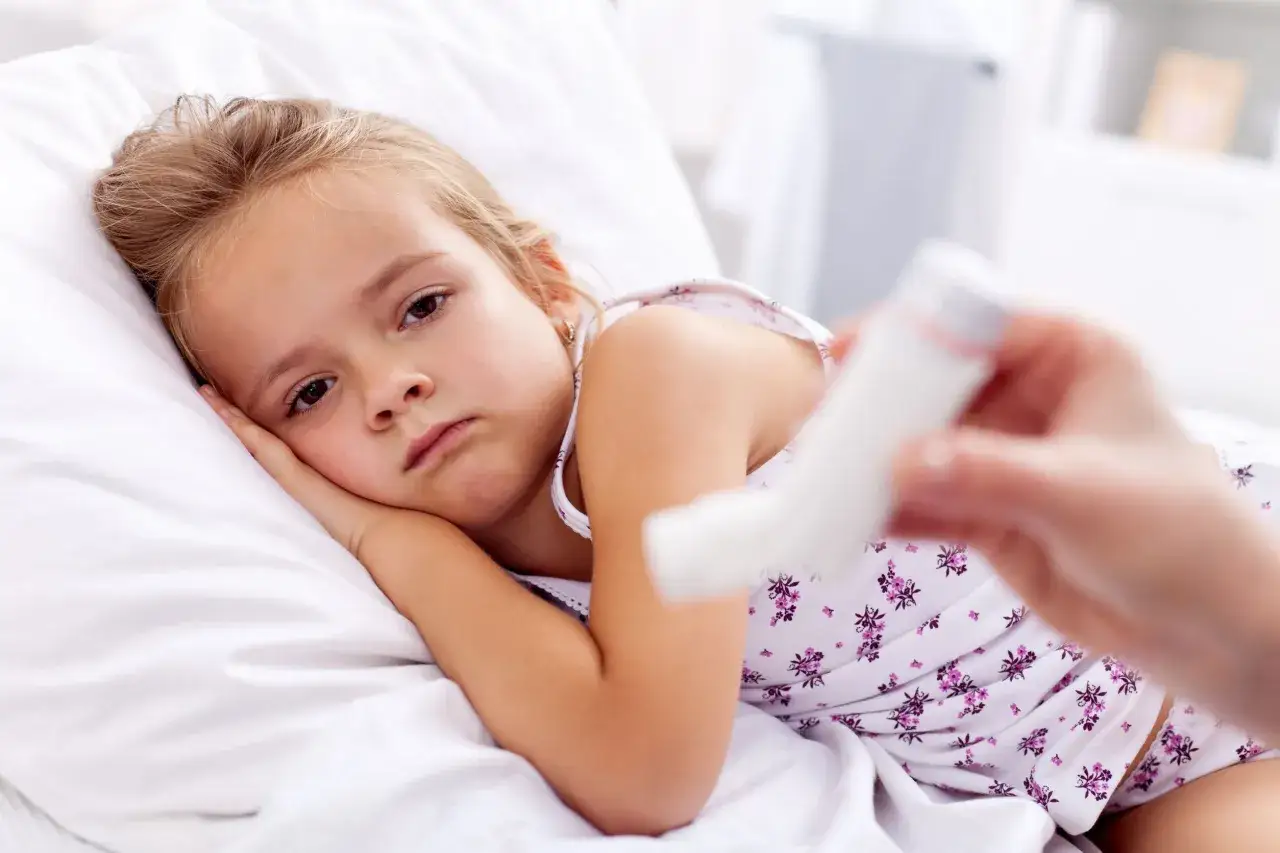 Smutna dziewczynka leży w łóżku, a ręka podaje jej inhalator. Dostępne są inhalacje dla niemowlaka.